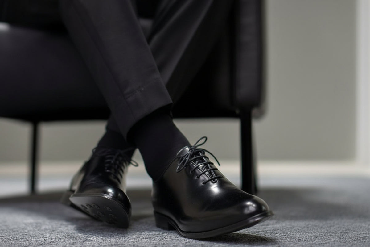 A close-up of a person's legs crossed, wearing black dress pants, black socks, and shiny black leather oxford dress shoes on a gray carpeted floor.