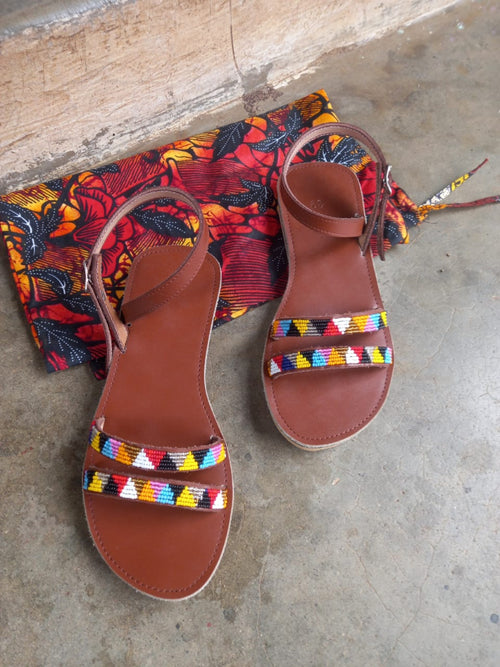 A pair of brown leather flat sandals with colorful geometric patterned straps, displayed next to a matching multicolor patterned drawstring pouch on a concrete floor.